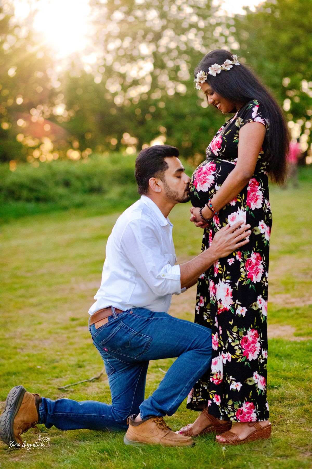 Husband kneeling to kiss his pregnant wife's bump in a sunlit park